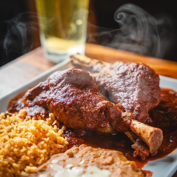 Steaming lamb shank in red sauce with rice and refried beans on a white plate, with a beer in the background.