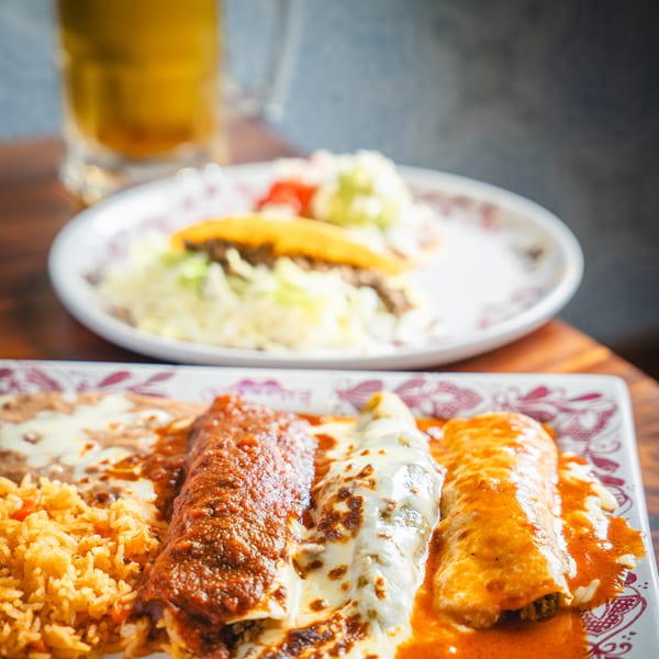 Three enchiladas with rice and beans on a patterned plate, with a taco and beer in the background.