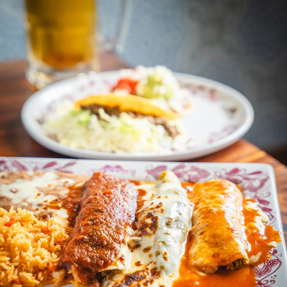 Three enchiladas with rice and beans on a patterned plate, with a taco and beer in the background.