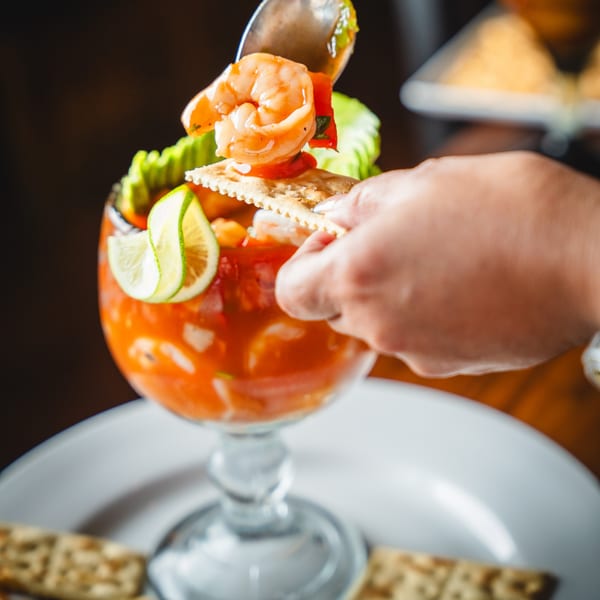 A hand spoons shrimp and sauce from a vibrant shrimp cocktail with lime and crackers.
