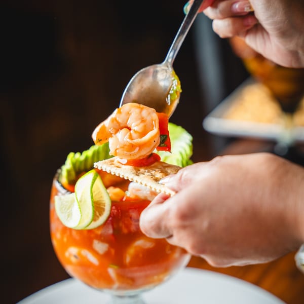 A hand uses a spoon to serve shrimp and sauce from a glass onto a cracker.