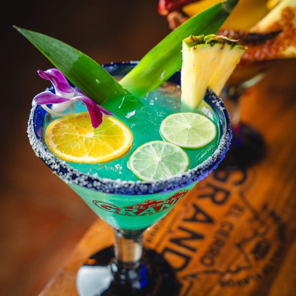 A vibrant blue margarita garnished with lime, orange, pineapple, a leaf, and an orchid.