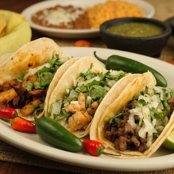 Three El Cerro Grande tacos, garnished with cilantro and onions, alongside peppers, rice, and beans.