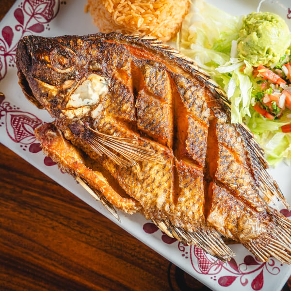 Crispy fried whole fish with rice, guacamole, and salad on a decorative white plate.