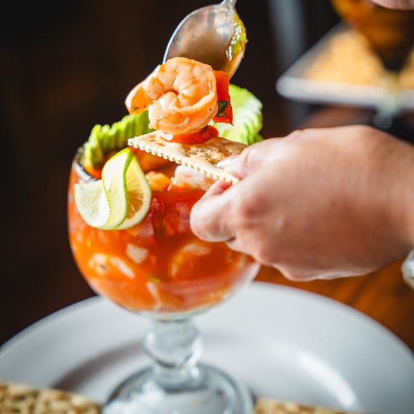 A hand spoons shrimp and sauce from a large glass of ceviche with crackers and lime.