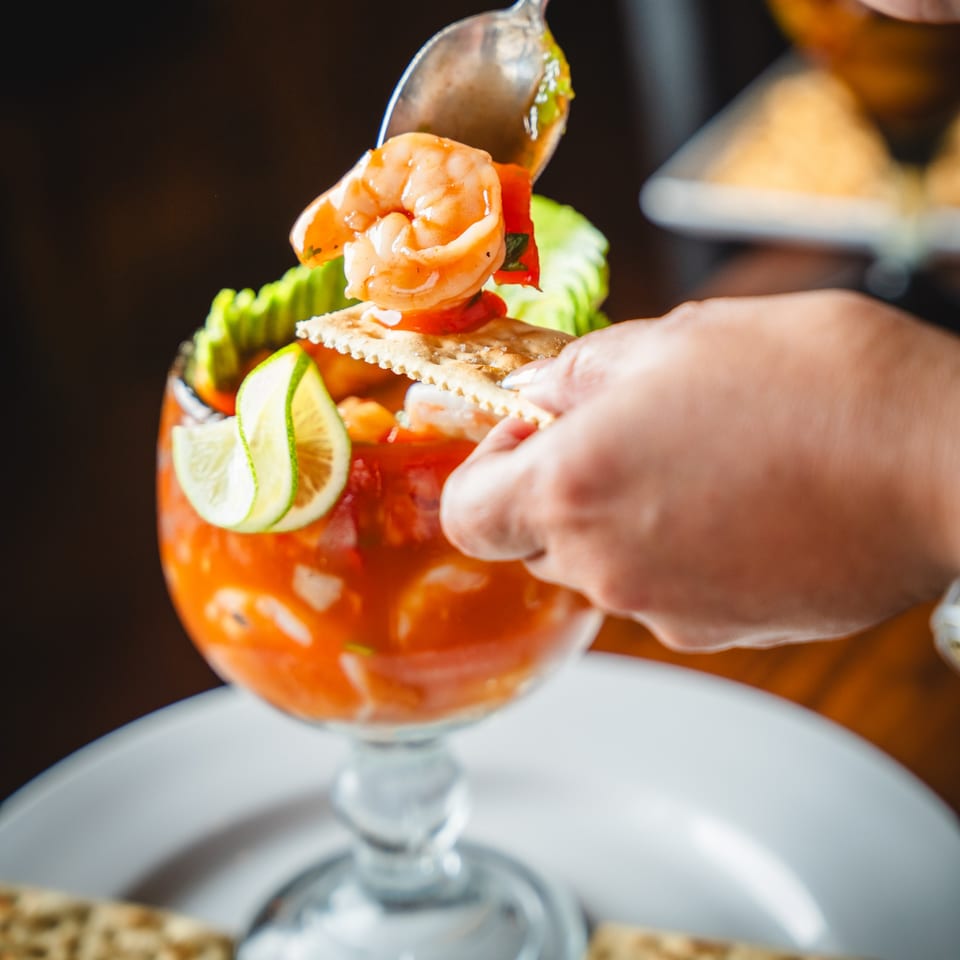 A hand spoons shrimp and sauce from a large glass of ceviche with crackers and lime.