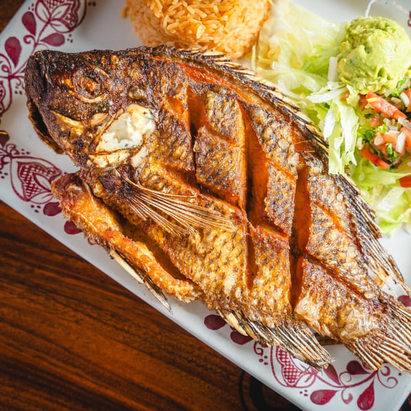 Crispy fried whole fish with rice, guacamole, and salad on a white plate with red patterns.