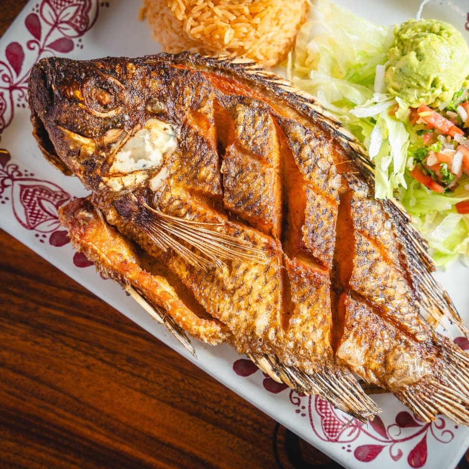 Crispy fried whole fish with rice, guacamole, and salad on a white plate with red patterns.