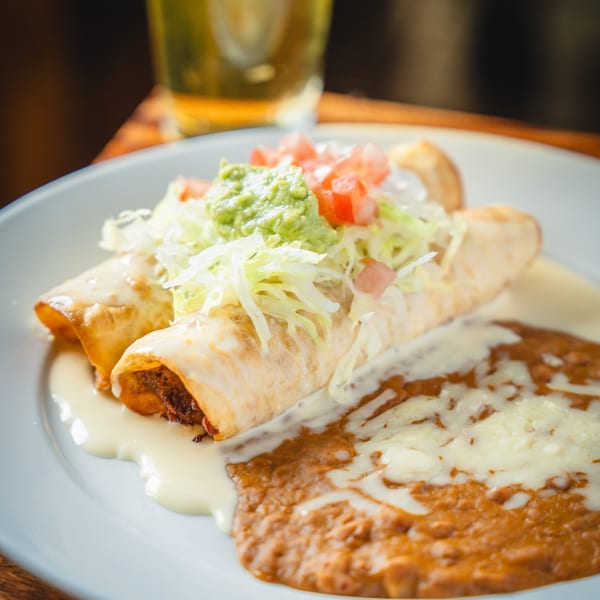 Two golden-brown enchiladas topped with guacamole, diced tomatoes, and shredded lettuce, alongside refried beans.