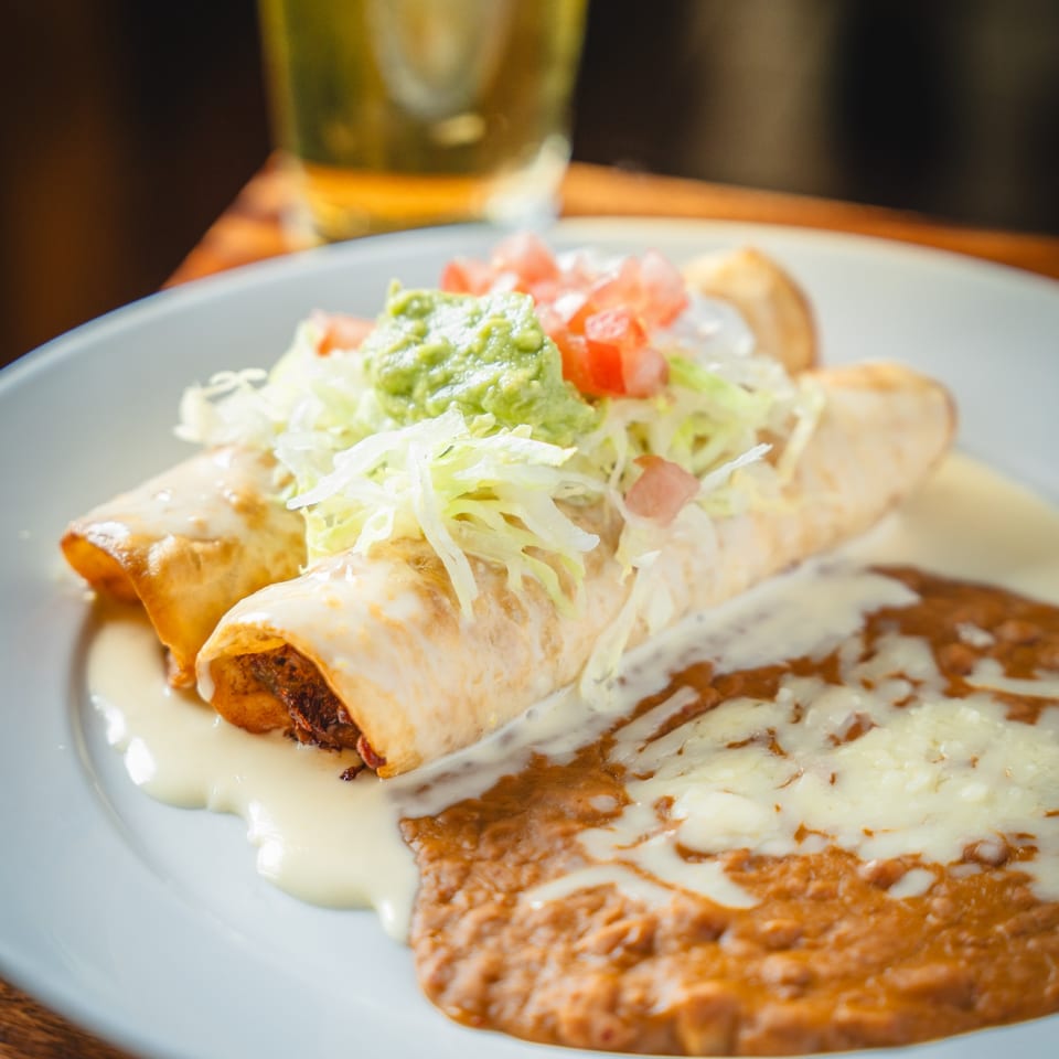 Two golden-brown enchiladas topped with guacamole, diced tomatoes, and shredded lettuce, alongside refried beans.