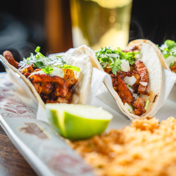 Three steaming tacos with meat, cilantro, and onions, served with lime and rice.