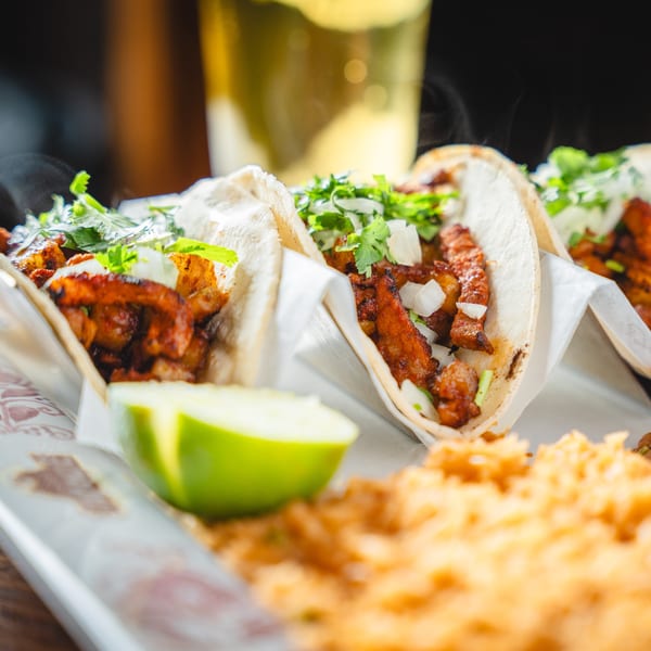 Three steaming tacos al pastor with cilantro, onions, and a lime wedge, served with rice.