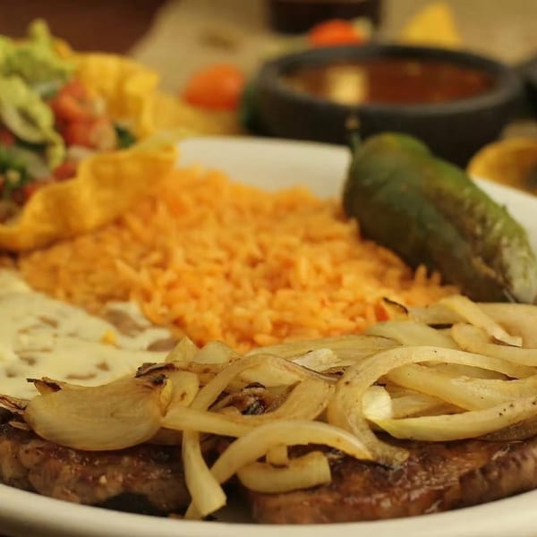 A close-up of a Mexican dish with steak, grilled onions, rice, and a green pepper.