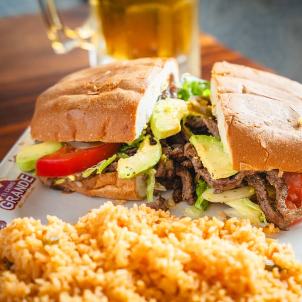 A large El Cerro Grande torta with steak, avocado, lettuce, and tomato, served with rice and a beer.