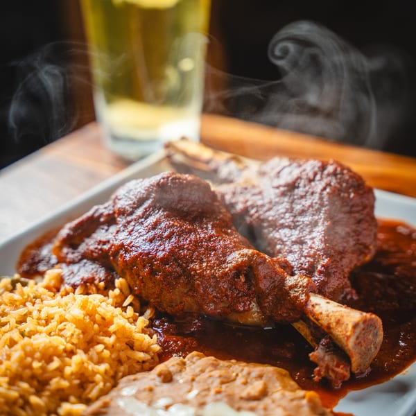 Steaming lamb shanks in rich red sauce, with rice, refried beans, and a beer.