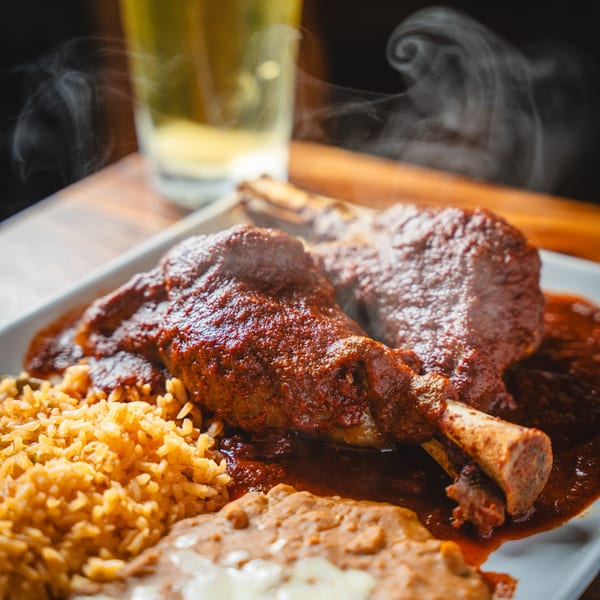 Steaming lamb shanks in rich red sauce with rice and refried beans, a beer in the background.
