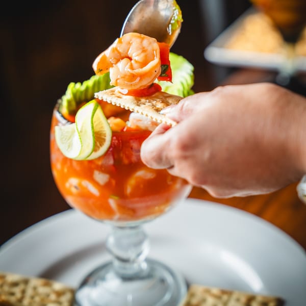 A hand uses a spoon to scoop shrimp and sauce onto a cracker from a large glass of ceviche.