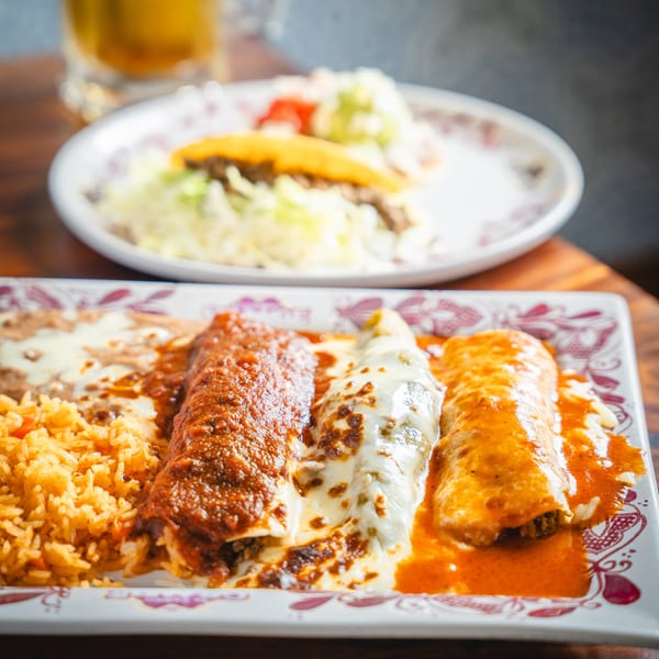 A plate of three enchiladas with rice and refried beans, and a taco in the background.