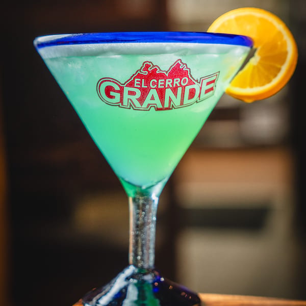 A vibrant green margarita in a branded El Cerro Grande glass, garnished with an orange slice.