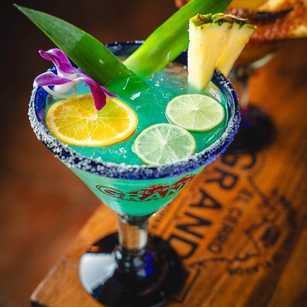 A vibrant blue margarita with lime, orange, pineapple, and an orchid, on a wooden surface.