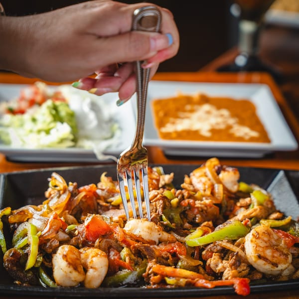 A hand uses a fork to pick up a shrimp from a sizzling plate of fajitas with sides.