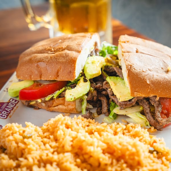 A steak torta with avocado, lettuce, and tomato, served with a side of rice and a beer.