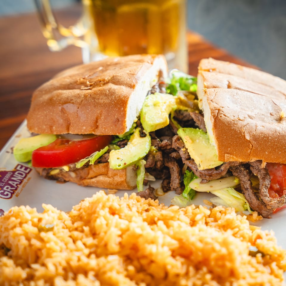 A steak torta with avocado, lettuce, and tomato, served with a side of rice and a beer.