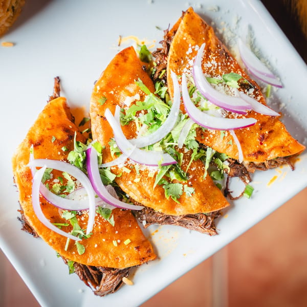 Three birria tacos with shredded beef, cilantro, and red onion on a white rectangular plate.