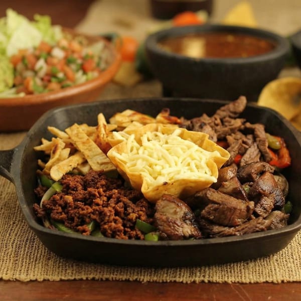 Sizzling fajita platter with steak, ground beef, cheese, and tortilla strips, with salsa and salad.