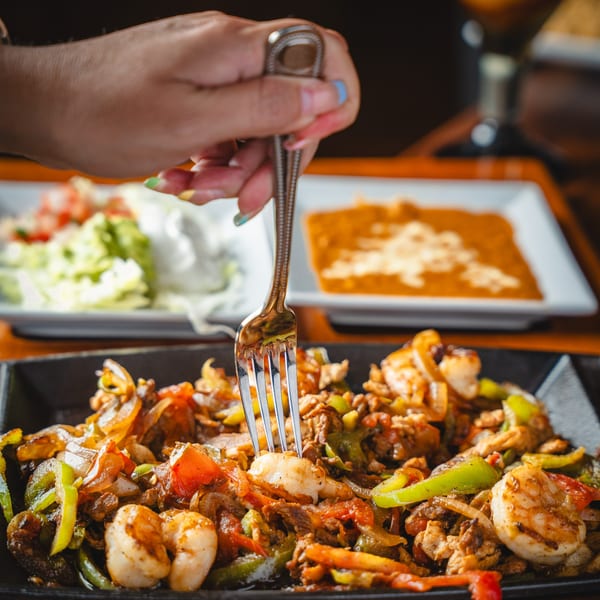 A hand with blue nail polish uses a fork to pick up a shrimp from sizzling fajitas.