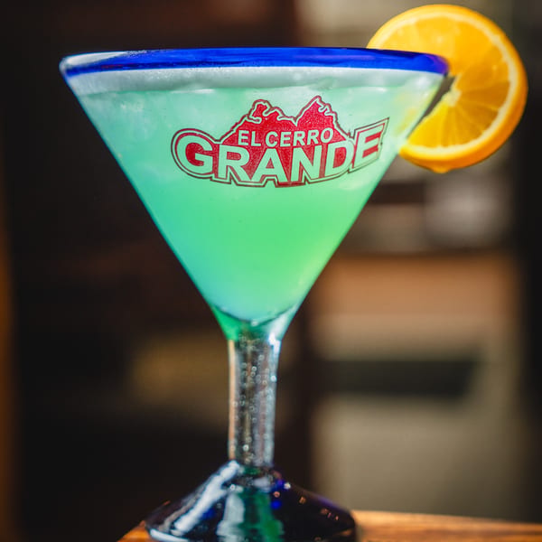 A bright green El Cerro Grande margarita in a blue-rimmed glass with an orange slice.