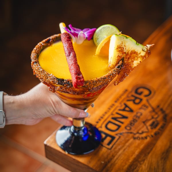 A hand holds a mango margarita garnished with a chili straw, lime, pineapple, and orchid.
