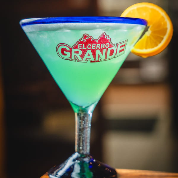A green margarita in a branded El Cerro Grande glass with a blue rim and orange slice.